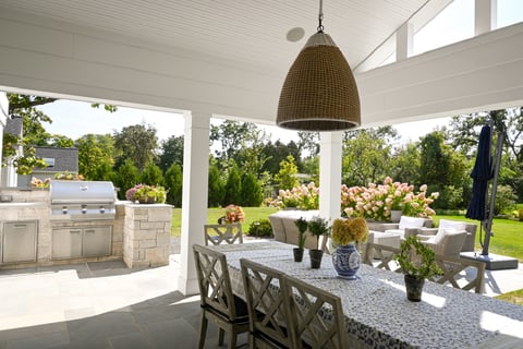 Residential backyard with a white pergola and prepared dining table with lighting and chairs on stone patio